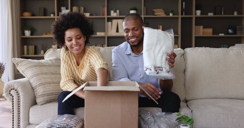 African couple sit on sofa at home unpack parcel box, take out bought online fashion winter clothes. Happy satisfied clients of e-commerce retails services. Shoppers family, remote e-shopping concept