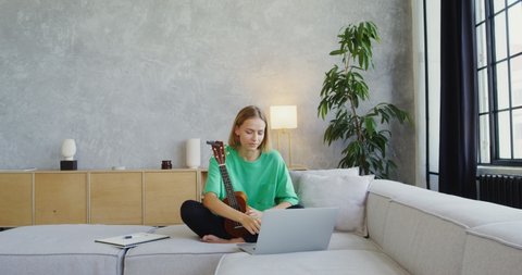 A young woman of European appearance plays the ukulele while video calling on a laptop, sitting on a sofa in a modern light interior. Online learning to play a musical instrument