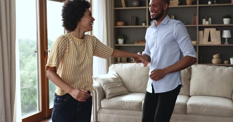 Merry African 30s couple in love dancing in living room, holds hands enjoy music spend weekend at home. Happy homeowners family celebrate relocation day. Hobby, bank loan, new property owners concept