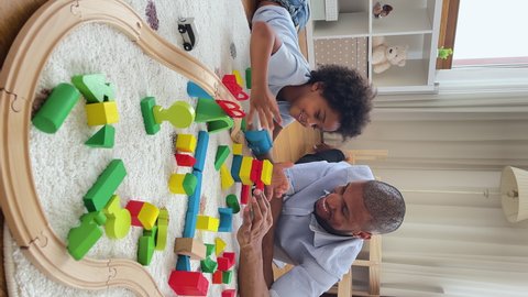 Vertical view, loving African dad play wooden cubes with 6s son, lying on warm floor in cozy nursery enjoy modern playthings, build tower from blocks and communicate. Family games and leisure concept