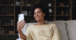 Mixed race teen female spend free time for making videocall on modern cellphone shoot herself in video clip. Smiling young afro american lady sit on sofa wave hand to phone webcam starting stream live