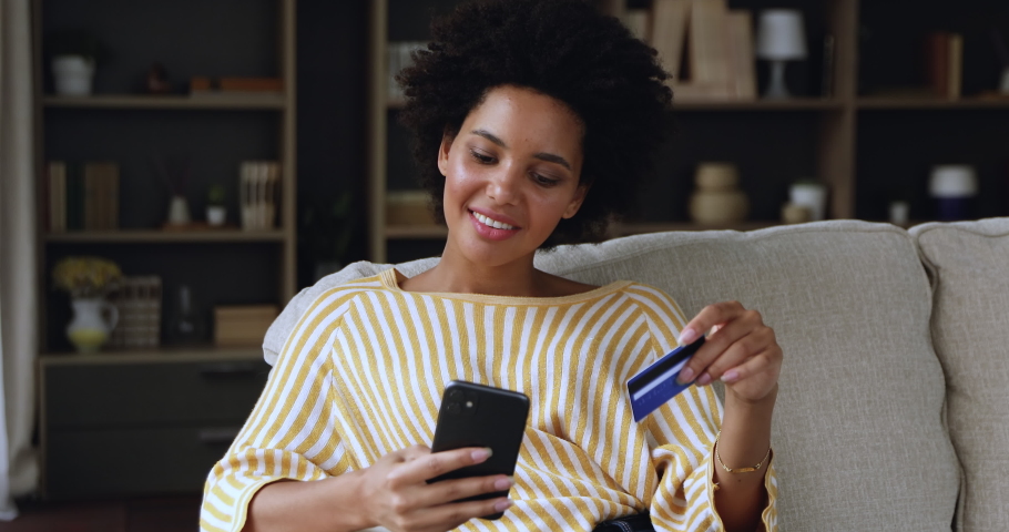 Satisfied gen z mixed race lady credit card holder use phone to pay online buy consumer goods order services on ecommerce websites from home. Happy young black woman make safe transaction at ebank app Royalty-Free Stock Footage #1079732981