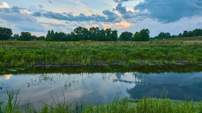 A small quiet river with overgrown banks. Summer countryside. Picturesque warm nature. In the distance is a village with wooden houses. Clouds float quickly across the sky.