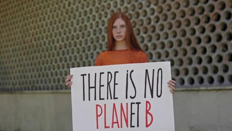 Young female activist with long red hair standing alone on street and holding cardboard with phrase there is no planet B. Caucasian teen girl protecting nature and environment.