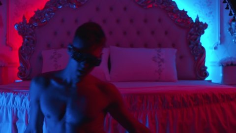 Young man stripper acting sexy and dancing on the floor in neon lighting - sits down on the bed and continue his dance