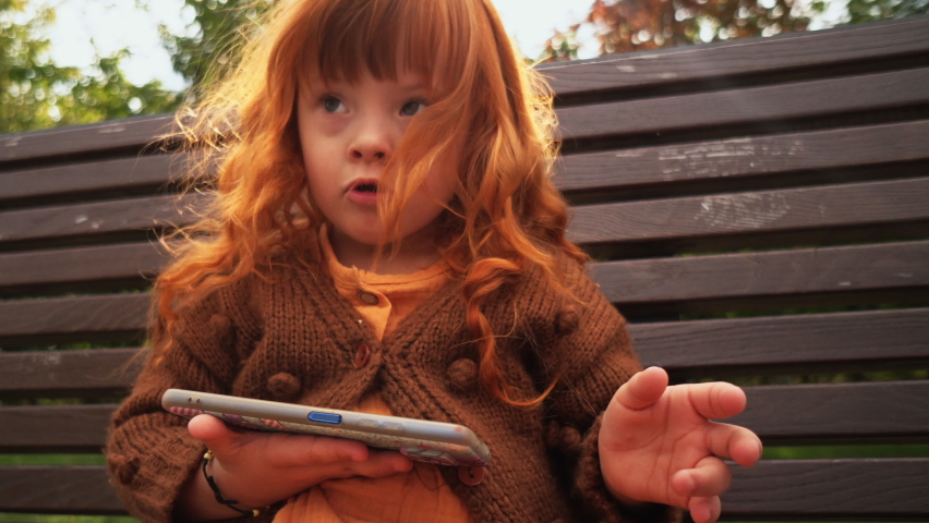 Toddler girl with down syndrome sitting on the bench and playing on smartphone | Shutterstock HD Video #1079736833