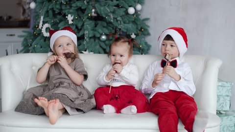 4k video portait of happy cute family of three children sitting on coach and eating holiday sweet xmas chocolate candies. Kids wearing Christmas red Santa hats and dressed in cute holiday clothes