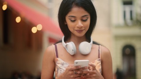 Portrait of attractive Indian girl with headphones on her neck texting on her phone. Beautiful young lady at the city street.