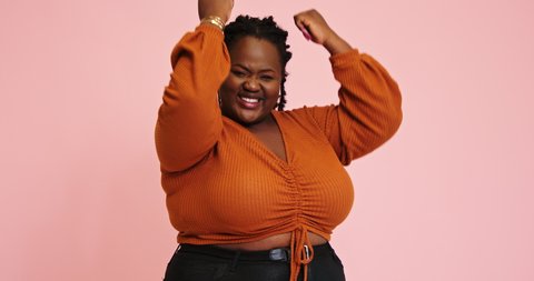 African-American plus size woman rejoices with winning and expresses emotions raising fists for camera against pink wall slow motion