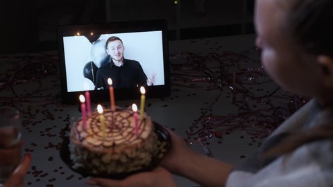 Online birthday party. Video congratulation. Distance celebration. Happy guy on tablet screen greeting with best wishes female friend blowing candles on cake.