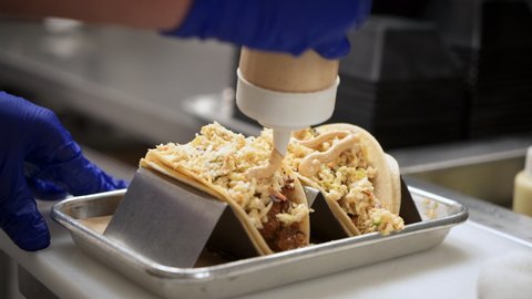 Chef drizzles sauce on top of fish tacos plated on metal tray taco stand rack, close up slow motion 4K