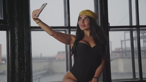 Beautiful young lady in black bodysuit and yellow kerchief making selfie photo on smartphone near window in house against urban cloudy cityscape. Tanned brunette girl with perfect sporty sexy back mov