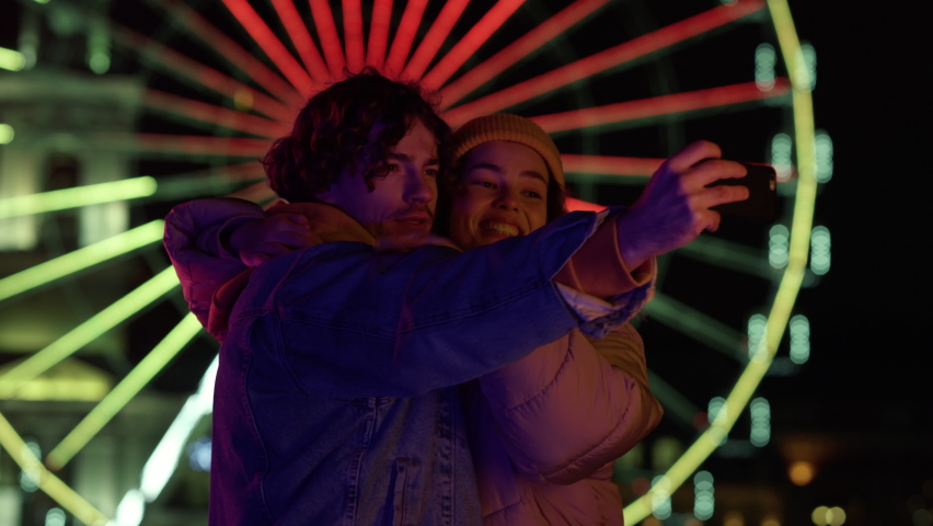 Smiling couple taking selfie photo on cellphone outdoor at night. Cheerful man and woman grimacing at mobile phone camera on ferris wheel lights. Joyful guy and girl using smartphone at night.  Royalty-Free Stock Footage #1079749391