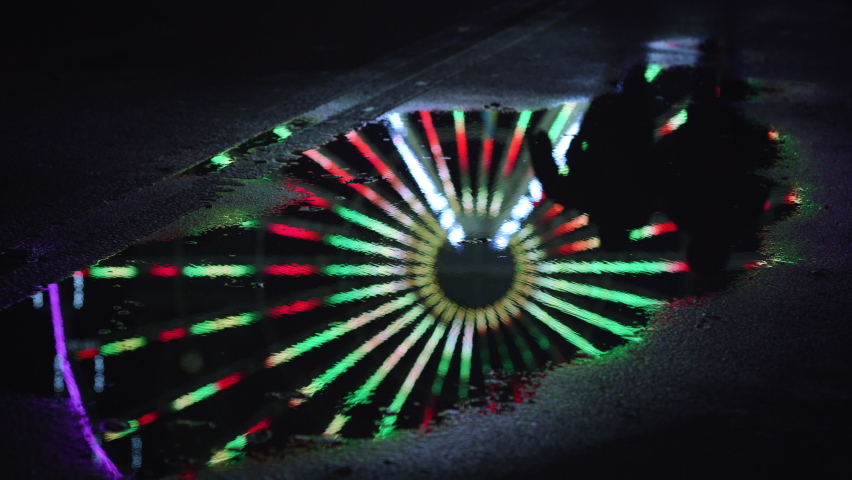 Reflection of unknown couple walking at puddle on urban street. Unrecognizable people feet running through water surface in city center. Mirroring of colorful illuminated ferris wheel.