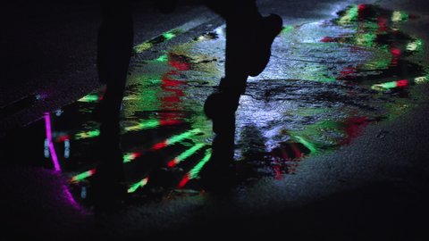 Reflection of unknown couple walking at puddle on urban street. Unrecognizable people feet running through water surface in city center. Mirroring of colorful illuminated ferris wheel.