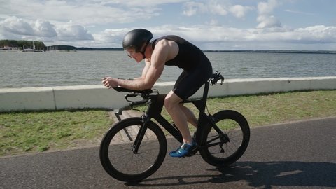 Cinamatic shot of Triathlete rides a cutting bike, pro cyclist rides on a track near a lake, athlete training for triathlon, 4k super slow motion.