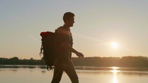 Silhouette of a young man hiking with a huge backpack along the river at sunrise. Guy enjoying his the journey. Victory over oneself. Concept of trekking, healthy lifestyle, travelling. Solo activity