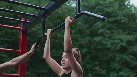 Beautiful Young Caucasian Female in a Ponytale Doing Pull Ups at an Outside Gym, Wearing a Black Sports Bra and Grey Pants, White Caucasian Male Personal Trainer Helping Her Out By Encouraging.