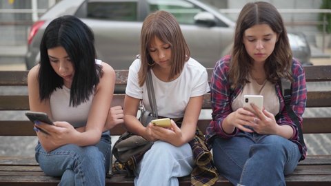 Front view portrait of three absorbed teenage girls surfing social media in smartphone apps sitting on city street. Adolescent Caucasian friends ignoring communication engrossed in Internet outdoors