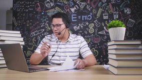 Asian man wearing glasses having a meeting online on laptop, taking note and using wireless microphone on the table at home, serious talk. Trying to explain work. Working at home concept