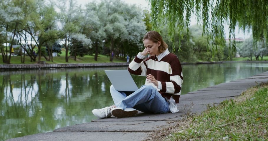 A beautiful young girl of European appearance uses a laptop sitting on the sidewalk on the embankment in the lotus position, enjoying the surrounding nature
