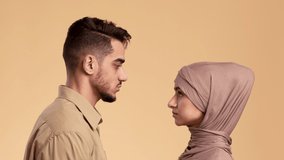 Serious Muslim Couple Turning Head And Looking At Camera Posing Over Beige Studio Background. Portrait Of Unhappy Ismalic Man And Woman Wearing Traditional Hijab Headscarf Concept. Slow Motion