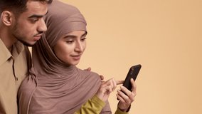 Excited Muslim Couple Using Smartphone Standing Over Beige Studio Background. Middle-Eastern Wife Showing Husband Great New Mobile Application For Cellphone. Cropped