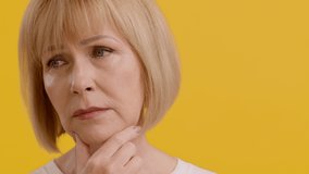 Closeup Portrait Of Pensive Senior Woman Touching Chin And Looking Up, Doubtful Middle Aged Lady Thinking About Something While Standing Isolated Over Yellow Studio Background, Slow Motion Footage