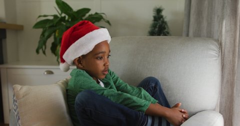 Sad african american boy wearing santa hat sitting on sofa at christmas time. 