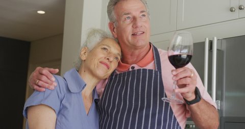 Happy caucasian senior couple wearing aprons embracing and drinking wine. 