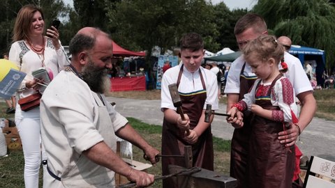 Ukraine, Vinnytsia Circa 2021. A blacksmith at a festival teaches a boy and a girl to forge an iron horseshoe with a hammer. Blacksmithing craft.