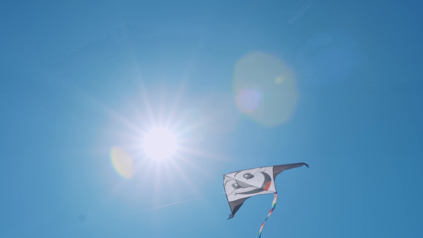 Colourful smiling flying rainbow kite on clear blue sky and sun rays background, kids flying toy for leisure activity. Flying rainbow kite with swaying tail in the air, children entertainment games