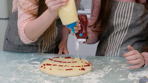 Close up hands of mother and daughter have fun with squeeze tomato sauce and mayonnaise on pizza dough during cooking together in home kitchen.