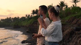 Young romantic couple in love standing near ocean on sandy shore in evening taking photo on mobile phone for share in social networks on holiday vacations, millennial female shooting video for vlog 