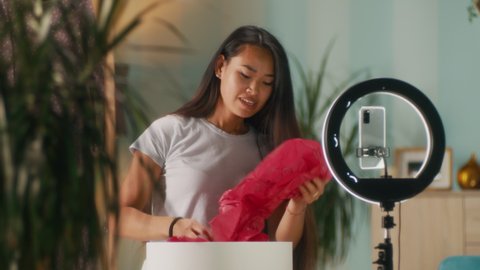 Happy Asian woman unpacking stylish dress from box and showing garment to audience while recording video for vlog at home