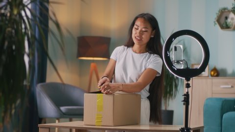 Friendly Asian woman in white t shirt opening carton box and speaking with audience during live stream at home