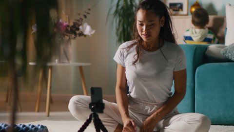 Asian woman in white clothes sitting cross legged near smartphone and showing warm up exercise to audience while recording video for fitness blog in living room with child