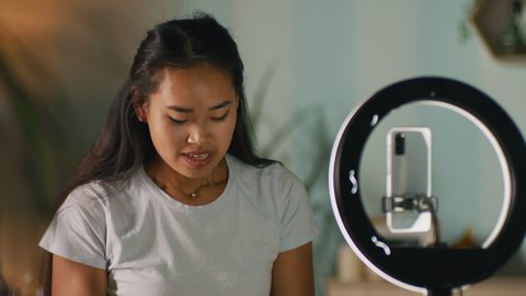 Young Asian woman looking at mirror and applying eye patches near child and ring light with smartphone while recording video for beauty vlog