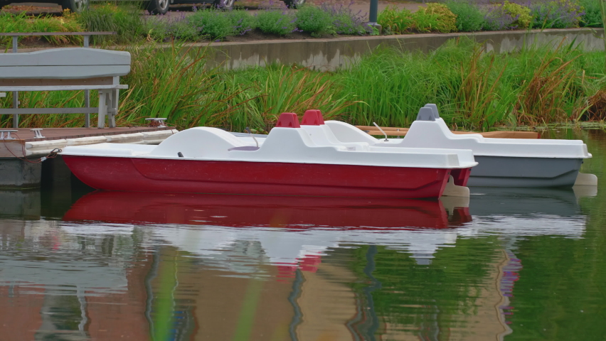 Red and gray small boats stand on the water at the pier, against the background of green grass, on the pier there is a gray bench