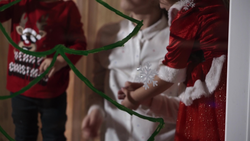 Authentic cute mom and two small preschool minor children siblings girl and boy 2-4 years old draw Christmas tree on window. young woman with children glues snowflakes. Xmas, New Year, winter concept.