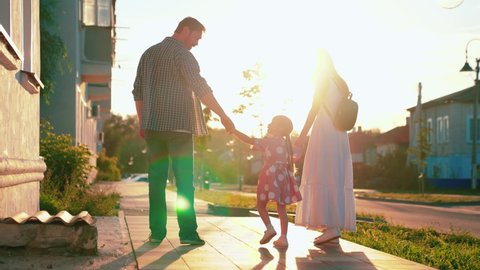 Happy family walks with child on quiet street in summer. Happy parents and children walking in park. Dad mom and daughter are holding hands on sidewalk in city in sun. Family vacation, weekend