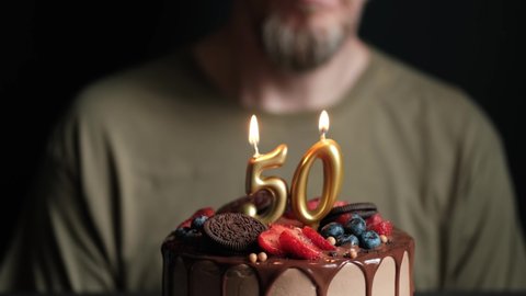 Fifty year old man blows out burning numbers 50 candles on chocolate cake and claps your hands. Birthday cake for 50 years anniversary. Slow Motion.