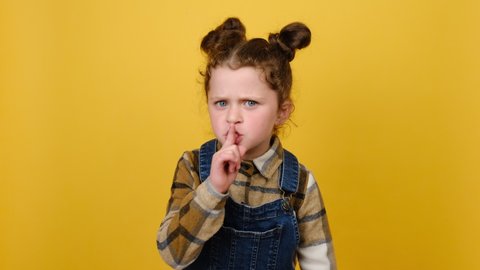 Secret little child girl saying hush be quiet with finger on lips shhh gesture close her mouth lock and throw away key, posing isolated over yellow color background studio. Childhood lifestyle concept