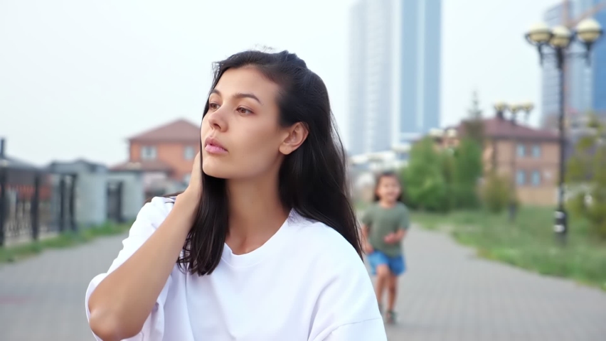 Playful Korean girl runs and jumps on thoughtful mother in white t-shirt back squatting on city embankment in summer evening slow motion closeup