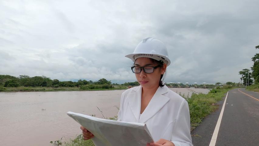 A female engineer holding a blueprint walks around the site of the river