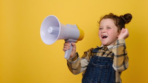 Portrait of cheerful funny little child girl screaming in megaphone clenching fist looking aside, posing isolated over yellow color background wall in studio. Childhood emotion lifestyle concept