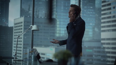 Confident Young Businessman in a Suit Standing in Modern Office, Talking on a Phone, Looking out of the Window on Big City with Skyscrapers. Successful Finance Manager Planning Work Projects.