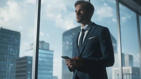 Confident Young Businessman in a Suit Walking in Modern Office, Using Smartphone, Looking out of the Window on Big City with Skyscrapers. Successful Finance Manager Planning Work Projects.