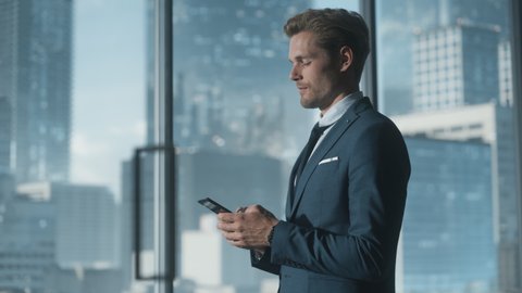 Confident Young Businessman in a Suit Standing in Modern Office, Using Smartphone, Looking out of the Window on Big City with Skyscrapers. Successful Finance Manager Planning Work Projects.
