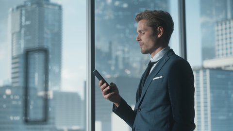 Confident Young Businessman in a Suit Standing in Modern Office, Using Smartphone, Looking out of the Window on Big City with Skyscrapers. Successful Finance Manager Planning Work Projects.